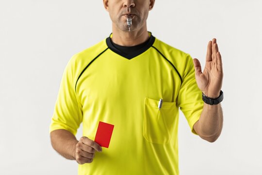 Professional referee in yellow jersey blowing a whistle while showing a red card, indicating expulsion and stopping play