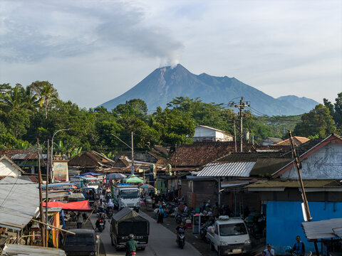 Klaten, Indonesia - 23 April 2026: Aerial view of Pasar Kembang Klaten traditional market with the smoking Mount Merapi volcano in the background, featuring busy street traffic and local rooftops.