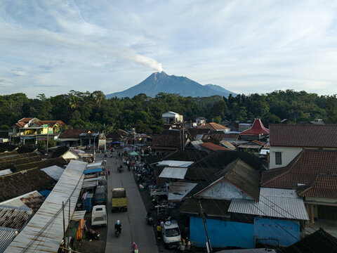 Klaten, Indonesia - 23 April 2026: Aerial view of Pasar Kembang Klaten traditional market with Mount Merapi emitting smoke in the background under a blue sky.