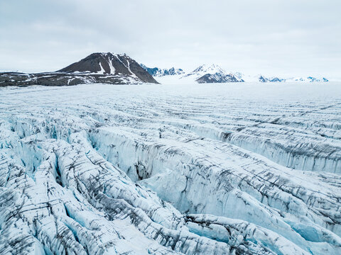 Svalbard and Jan Mayen - 24 June 2025: Aerial view of the vast crevassed surface of a glacier with blue ice and rugged mountains under a cloudy sky.