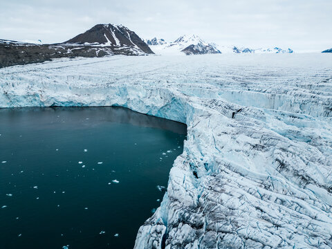 Svalbard and Jan Mayen - 24 June 2025: Aerial view of a massive glacier wall meeting the dark arctic sea with floating ice chunks and snow-capped mountains under an overcast sky.