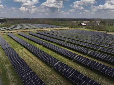 Lebanon, United States - 12 April 2026: Aerial view of a large solar farm with rows of photovoltaic panels on green grass under a cloudy sky with a red barn in the background.