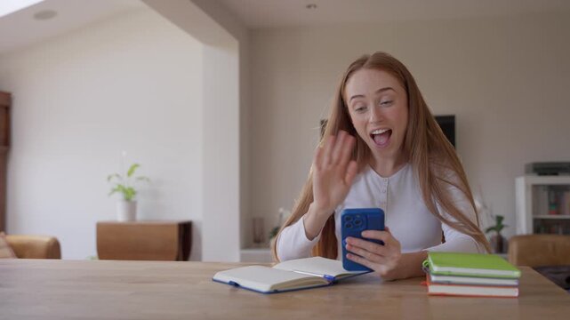 Girl sitting at a table looking at her phone and taking selfies while studying in a modern house living room