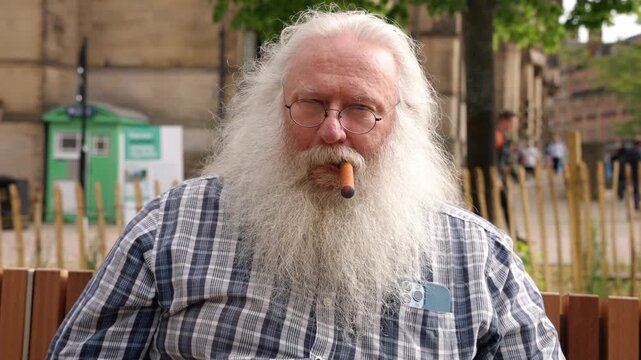 Elderly man smokes cigar while sitting outdoors with green trees and buildings in the background during daytime
