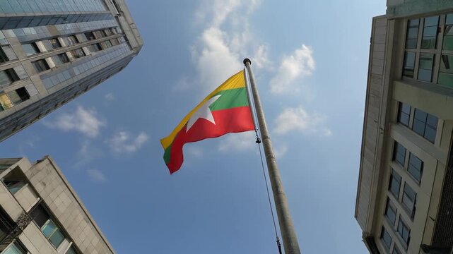 Stock image showing the national flag of Myanmar flying high on a pole with a city building corner under sunny sky.