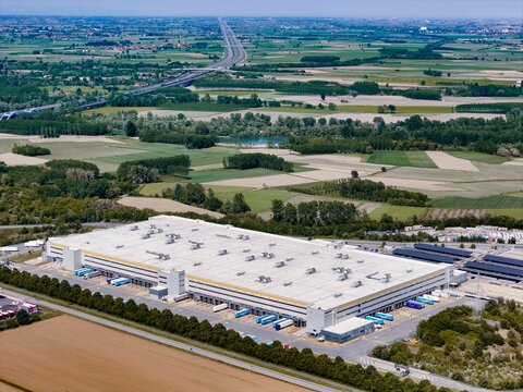 Torrazza Piemonte, Italy - 03 May 2026: Aerial view of the Amazon TRN1 fulfillment center with trucks at loading docks and surrounding green fields under a clear sky.