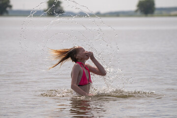 Naklejka premium Female swimmer in pink bikini splashes water while flipping hair back in a lake, with trees and a clear sky visible in the background