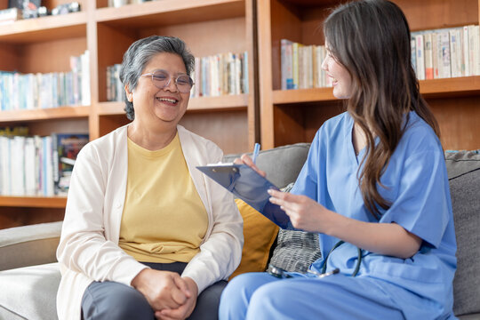 Happy smiling elderly woman patient sitting with nurse at home holding clipboard providing professional health care advice during friendly living room consultation near wooden bookshelf indoors