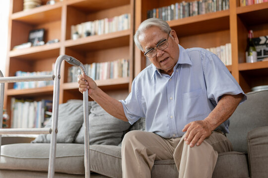 Asian elderly man sitting comfortable gray sofa holding aluminum walker showing pain facial expression feeling painful knee joint experiencing physical distress resting alone cozy living room