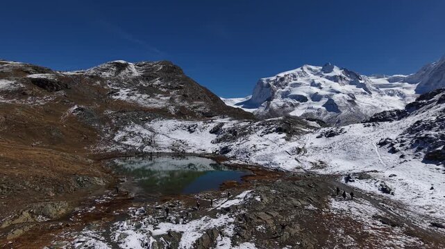 Aerial flyover above Riffelsee lake towards Gorner Glacier in Zermatt, Switzerland with the famous Monte Rosa and Dufourspitze peaks in the distance behind the ice on a autumn day in the Swiss Alps