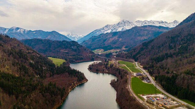Klaus an der Pyhrnbahn, Austria - 23 March 2026: Aerial view of the Klauser Stausee reservoir winding through a valley with snow-capped mountain peaks and the VOEST Quarry under a cloudy sky.