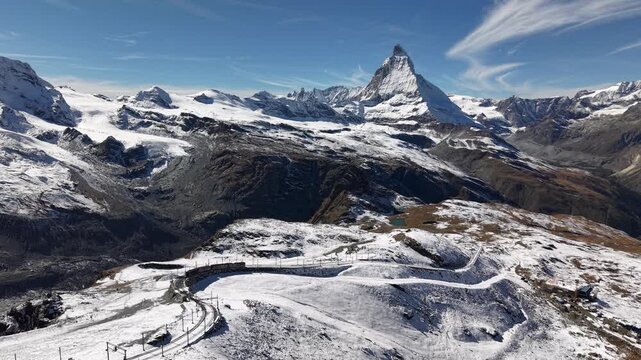 Aerial flyover over alongside the Gornergrat railway in Zermatt, Switzerland with a view of a train riding down the tracks and the Matterhorn on a snowy autumn day in the Swiss Alps