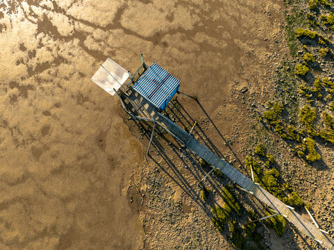 Yves, France - 22 September 2023: Aerial view of a traditional fishing hut on stilts with a square net and a long wooden walkway over the muddy tidal flats during golden hour.