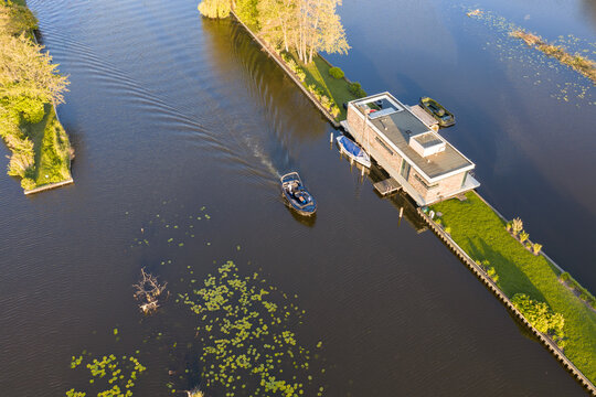 Breukelen, Netherlands - 25 April 2026: Aerial view of Scheendijk with a modern houseboat and a small boat cruising through the calm canal waters surrounded by lush green strips of land and lily pads.