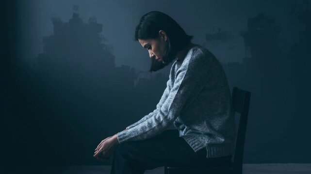 A woman sits alone on a chair in a dimly lit room deep in thought and emotional reflection