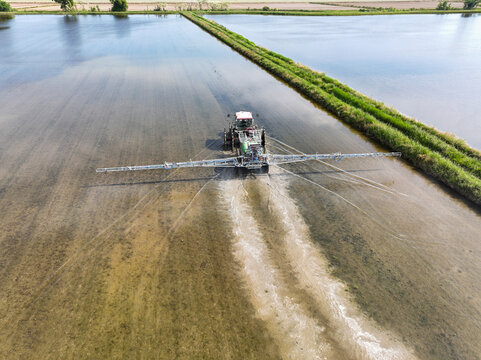 Aerial view of a tractor with a wide sprayer boom working in flooded rice paddies under a bright sky in Trino, Piedmont, Italy.