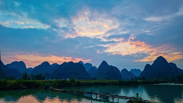 The calm stream reflects the fiery colors of the setting sun as a wooden bridge connects the fields, with lush greenery and majestic peaks completing this peaceful countryside scene.