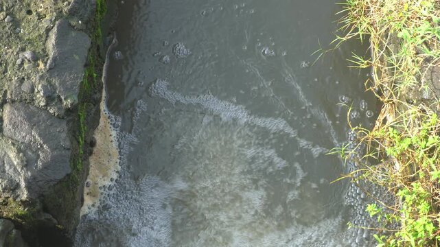 Grayish water flows slowly through a narrow rocky channel bordered by moss and grass. Foam and bubbles form on the surface.