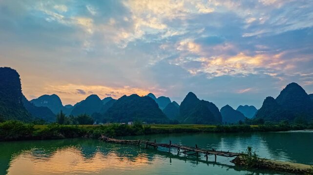 The calm stream reflects the fiery colors of the setting sun as a wooden bridge connects the fields, with lush greenery and majestic peaks completing this peaceful countryside scene.