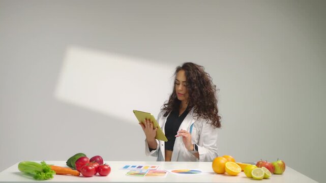 Female dietitian in white coat at desk with fruits, vegetables and colorful charts, using tablet to compose personalized meal plan. Healthy eating consultation and nutrition planning concept in studio