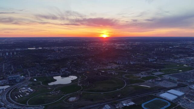 A cinematic aerial drone ascent over Downsview Park in North York, revealing the winding walking trails, commemorative pond, and vast suburban landscape under a fiery orange sunset
