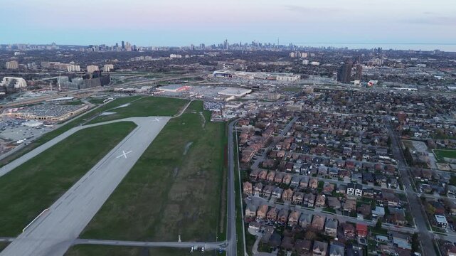 A slow lateral aerial drone slide left over North York, showcasing the closed runway of Downsview Airport, sprawling residential subdivisions, and the distant Toronto skyline at dusk
