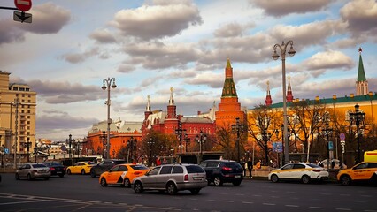 Moscow, Russia - April 22, 2026: Moscow Kremlin towers and city traffic at sunset. Vibrant urban scene featuring historical brick fortifications and yellow taxis on the road under a cloudy sky © Amina