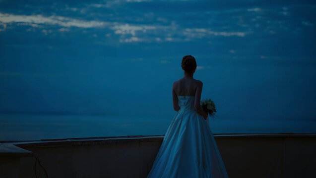 Solitary bride holding bouquet standing alone at night overlooking dark ocean horizon