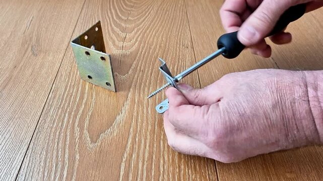 Close-up of hands using a screwdriver to tighten a screw through a metal bracket on a light wood surface. Detail of manual assembly, hardware tools, and DIY home repair project