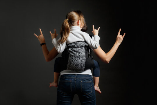 Mother and little girl in back carrier showing rock gestures with hands, studio shot from behind.