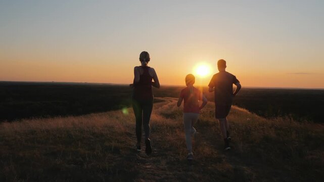 Active family jogging together along grassy hillside ridge at golden hour sunset.