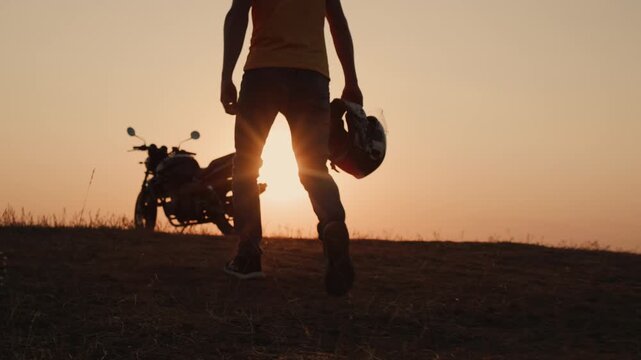 Silhouette of man holding helmet walking towards parked motorcycle during sunset.