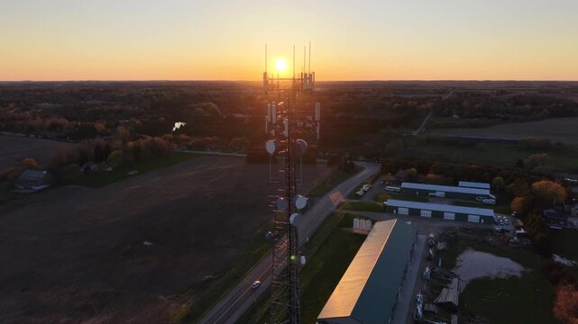 A cinematic aerial drone pull-back from a cellular communications tower at golden hour, revealing a winding country road, commercial outbuildings, and agricultural fields at sunset