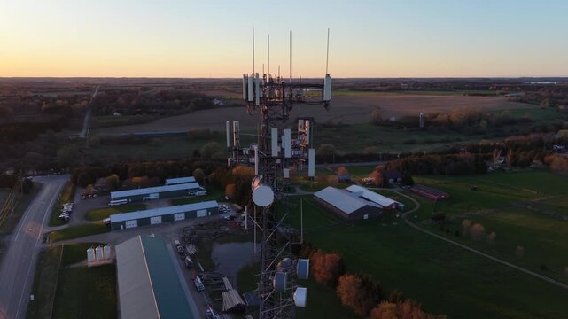 A cinematic 360-degree aerial drone orbit around a telecommunications tower at golden hour, capturing backlit microwave dishes and cellular antennas against a blazing sunset horizon