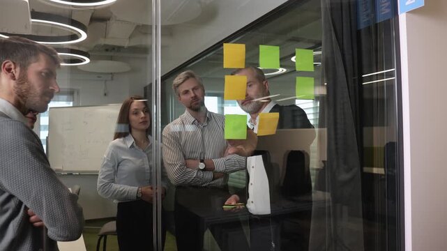 Businesspeople talking in their diverse team during a brainstorming session with sticky notes on glass wall in a modern office