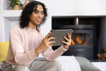 Naklejka premium Non-binary adult sitting on sofa in living room holding tablet, in pink sweater, fireplace burning