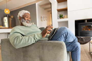 Naklejka premium Senior African American man sitting in gray armchair smiling while using tablet in cozy living room