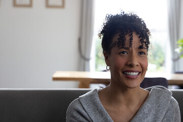 African American woman sitting and smiling on gray sofa in living room, showcasing hoop earring © wavebreak3
