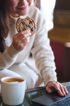 Vertical closeup image of a woman eating a piece of chocolate chip cookie while working on laptop computer in cafe