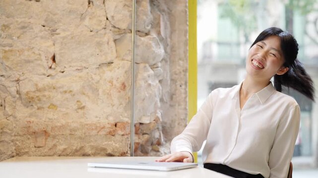 Young woman smiles, relaxed at a cafe table. She gestures gently while seated beside a laptop. Stone wall and glass window frame the scene. Natural light illuminates her calm, happy expression