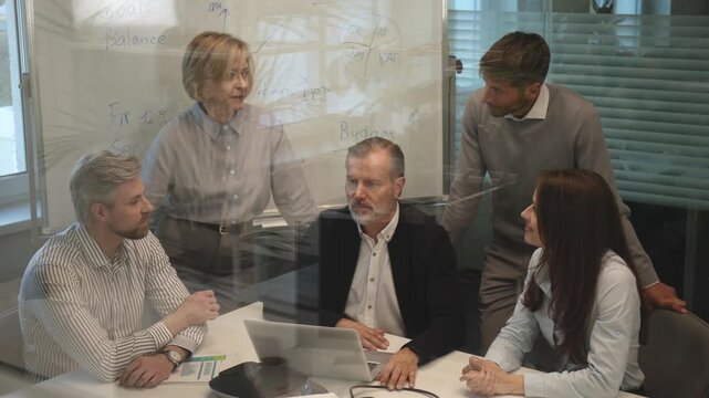 Successful businesspeople working in a coworking space working with laptop discussing strategy and business plan in modern office, view from glass wall