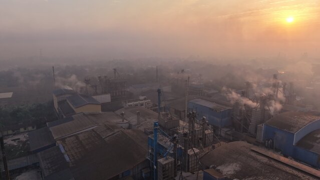 Aerial view of a large industrial sugar mill complex at sunrise with smoke billowing from chimneys into a hazy morning sky.
