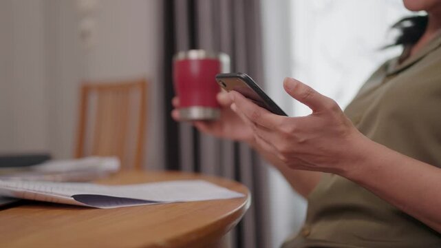 Smartphone Coffee Break A person engrossed in their smartphone, coffee in hand, sits by a table alongside paperwork, capturing a moment of technology-infused rest.