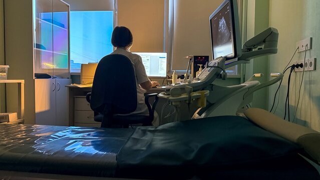 Female medical technician in ultrasound exam room, seated at imaging workstation with monitors and control panel, calm clinical mood, healthcare diagnostics setting