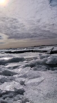 Frozen shoreline dusk with cracked ice, vast icy expanse with melting floes and shallow pools, low sun reflection on wet surfaces,