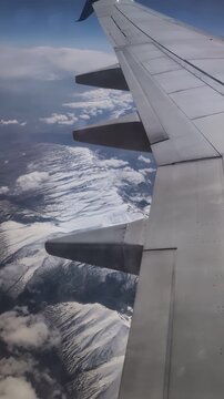 Wing over snowcovered mountain range at cruising altitude silver aircraft panel reflecting sunlight, soft cloud shadows