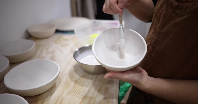 Talented artisan carefully applying a layer of glaze with a brush to a handmade white ceramic bowl in a pottery studio, preparing the piece for firing in the kiln