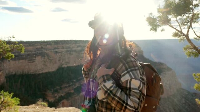 Young smiling woman with a backpack and a vintage camera hiking on the edge of the cliff at grand canyon national park, admiring the beautiful landscape with the sun flare in the background