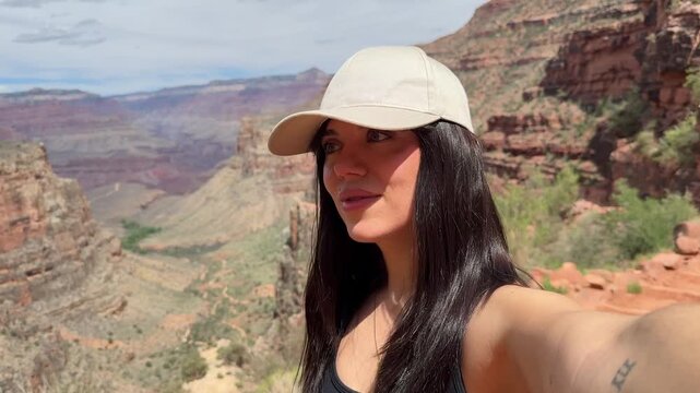 Beautiful young woman wearing a baseball cap filming a selfie video while hiking along the stunning bright angel trail with the magnificent grand canyon landscape in the background