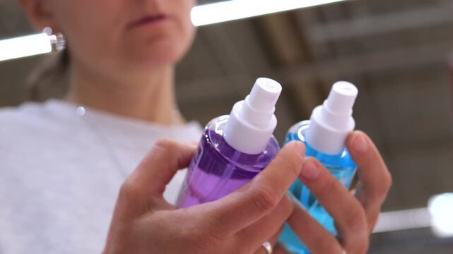 Focused customer holding two different cosmetic spray bottles in her hands, carefully comparing the purple and blue liquids while making a choice in a department store or supermarket